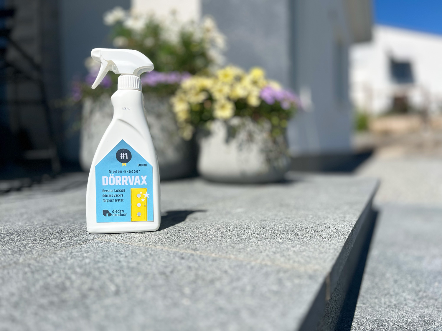 Sprayflaska med Dörrvax från Dieden Ekodoor, ett tillbehör för ytterdörrar, placerad på en trappa i sten framför en modern villa med blomkrukor i bakgrunden.