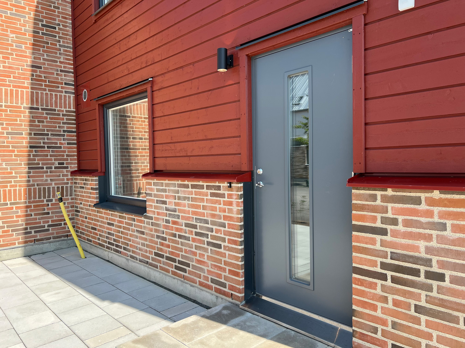 IMG_9360_komprimerad Grå ytterdörr med smalt glasparti från Dieden-Ekodoor monterad på fasad i rött trä och tegel